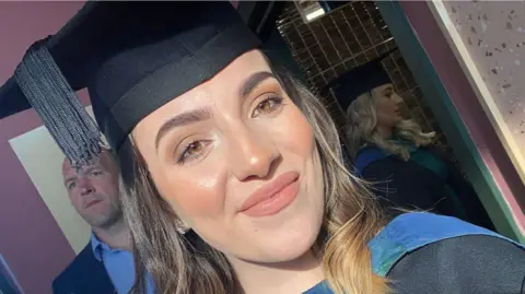 Sophie Morgan wears a black university cap and gown and smiles at the camera. She has brown short hair and brown eyes. 
