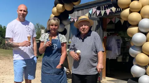 Three generations of a family - a younger man, a middle-aged woman and and elderly man - stand in front of an ice cream shop holding cones. Behind them the shop is heavily decorated with a balloon arch and bunting. 