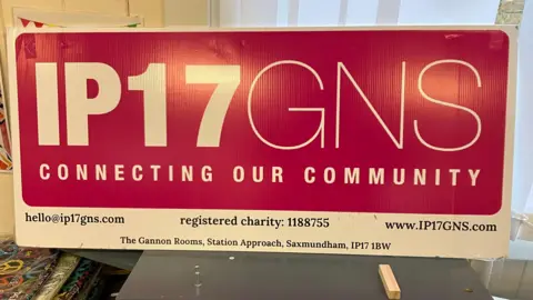 GUY CAMPBELL/BBC A red sign reading "IP17 GNS Connecting Our Community" is leaning against a window