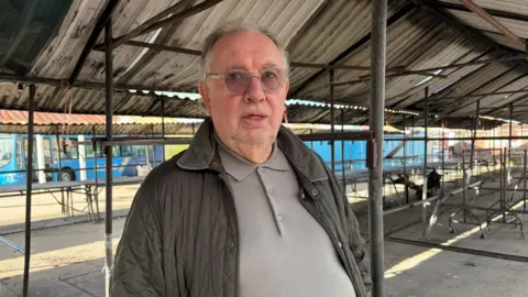 A middle aged man in a buttoned up grey shirt and jacket stands alone underneath metal clad roofing outside