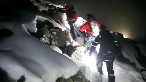 The members of Kintail Mountain Rescue Team are dressed in mountain attire and are walking through deep snow.