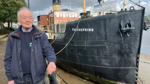 John Puddy, Chairman of the Freshspring Trust, standing in front of SS Freshspring moored up in Bideford Port.