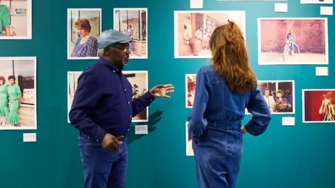 Two people dressed in denim stand in front of a blue wall featuring a number of frame photos
