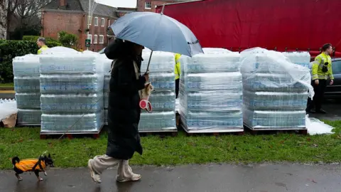 Getty Images A woman with am umbrella walking her dog by crates of bottled water.