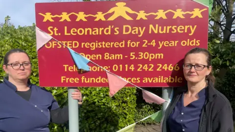 BBC/Simon Thake Two women stand either side of a crimson sign wearing blue uniforms. They look upset.