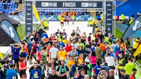Reading Borough Council Dozens of runners gather after crossing the finish line. A clock above the finish line displays the time 2 hours, 12 minutes and 37 seconds. In the foreground a marker flag say 2 hours and two runners in navy blue t-shirts take a selfie 
