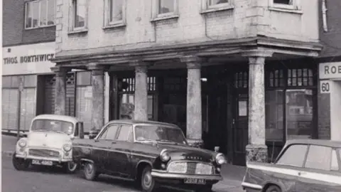 A black and white image of Kingsley Hall as seen from the street in the 1960s. The brick building's upper floors jut out over a pavement below and are supported by five columns. Three old-fashioned cars are parked just outside the building.