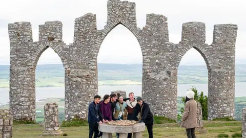 A large stone folly built overlooking a body of water, a group of people are huddled below it.
