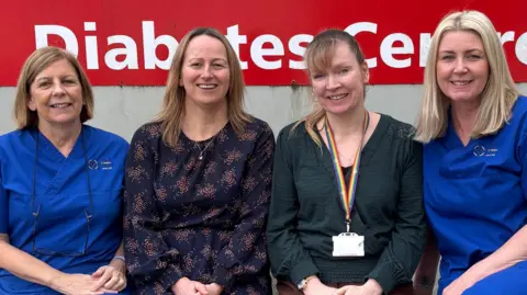 Hywel Dda University Health Board Four women sitting in front of a red and white hospital sign. Two are wearing blue hospital scrubs. They are smiling at the camera.