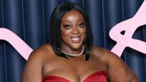 Judi Love smiles while wearing a red dress. She is wearing a silver necklace and has a tattoo on her left shoulder.