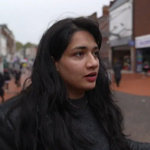 A woman with long dark hair wearing a dark grey jumper stands on a high street, looking to the right of the camera. The background is blurred but shops are visible on either side of the street.