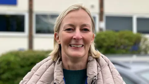 Lewis Adams/BBC Acting Detective Inspector Karen Osborne is wearing a beige coat in front of Harlow police station. She has short blonde hair and is smiling.