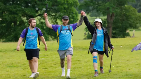 Family handout Ms Britton on the right with her brother Harry and partner Lloyd on the 26-mile fundraising walk in the Lake District. They are all wearing blue Alzheimer's Society t-shirts. shorts and walking shoes with trees in the background.