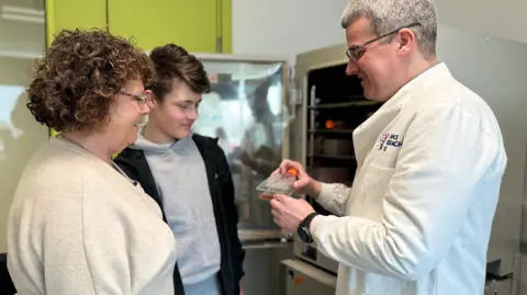 Julie and Jack are standing to the left of the shot and looking at one of the researchers in the lab, who is showing them a small piece of equipment.