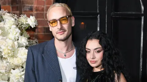 Getty Images George Daniel and Charli XCX stand next to one another and alongside a display of white flowers.