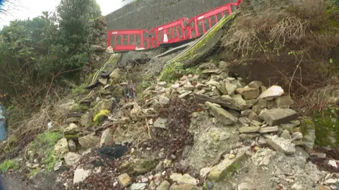 There is fallen rubble from the collapsed wall and red barriers at the top of the road. 