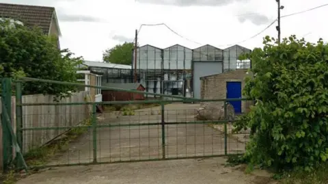 Google The entrance to Ravensworth Nurseries enclosed by a green metal gate in the foreground. The gate is slightly weathered and has horizontal and vertical bars forming a grid pattern. On either side of the entrance, there are fences and hedges, with a wooden fence visible on the left and a dense green bush on the right.
Beyond the gate, a concrete driveway leads into the yard, which appears somewhat worn and slightly uneven. To the right side inside the yard, there is a small brick outbuilding with a bright blue door. Nearby, there are a few scattered objects, including what looks like a wheel or round equipment piece leaning against the wall.