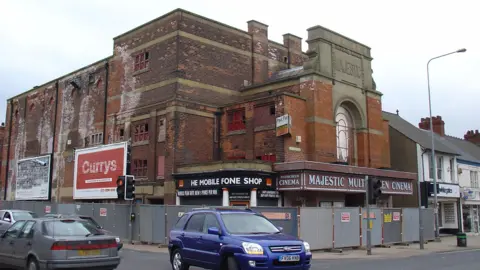 North Lincolnshire Museum The former Majestic Cinema in Scunthorpe made of brick and surrounded by construction fencing awaiting demolition. There are four cars near it.