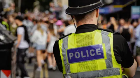 A police officer wearing a hi-vis jacket. He is stood in front of a crowd of people.