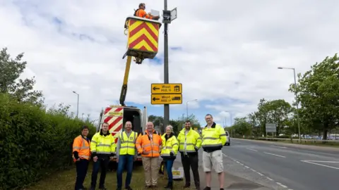 Seven people standing in a line wearing yellow and orange high-vis jackets and looking at the camera. They are standing on a grass verge next to a road. A highways van is behind them with a crane, and a man standing in the tub at the top.