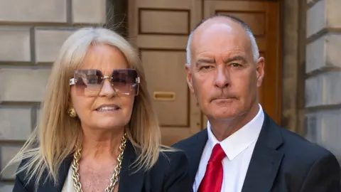 Tommy Sheridan with wife Gail standing outside the Court of Session - he is dressed in a black jacket, white shirt and red tie. He is mostly bald with white hair. She is wearing glam large sunglasses and gold jewellery