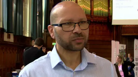 A man with short hair and black glasses in a blue striped shirt.