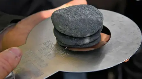Getty Images Shows a close-up of a hand holding a metal measuring plate with three flat stones stacked on it, with engraved text visible on the plate.