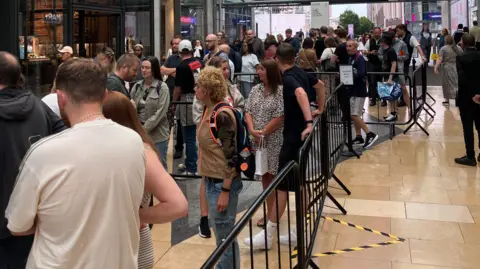 Men and women are queuing up near railings outside the Oasis Live '25 pop-up shop. A sign directs people where to queue. The Bullring shopping centre can be seen in the background.