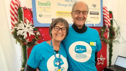 A man and woman smile with their arms round each other, both wearing blue t-shirt