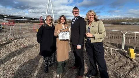 Four people standing on a building site. In the middle is Kim McGuinness in a light brown coat and standing next to her is Michael Mordey in a black suit and suit. He is wearing glasses. McGuinness is holding a clapperboard. They are all smiling. There is a white fence behind them. The sky is blue. 