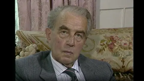 A screenshot from an archive interview with Francis Coulson. It is a head-and-shoulder portrait of Mr Coulson inside Sharrow Bay. His grey hair is combed back and he is wearing a grey suit, with a white and blue striped shirt and a grey and pink striped tie.