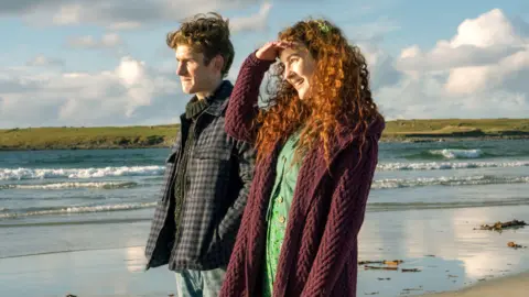 Vertigo Releasing Fionn O'Shea who is wearing a checked coat and Ann Skelly who wears a green skirt and shirt with a burgundy cardigan are standing on a beach and the sun shines on their faces.