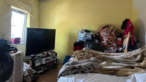 A room with yellow walls and mould on the ceiling, there is a bed and next to that there are bags piled up with items. There is a TV in the room.