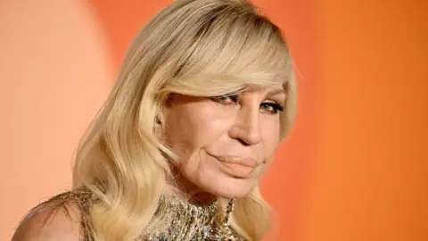 Getty Images Close-up dari Donatella Versace dengan gaun emas dengan latar belakang oranye.