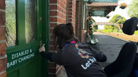 A young woman in a wheelchair is attempting to open a door on a train platform. She is wearing a grey t-shirt with the words "let me in" on the back. The door she is trying to open is green and says "disabled and baby changing".
