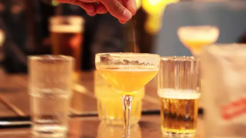 Getty Images Image of a bar with six drinks on it. The drink in the foreground is a yellow cocktail in a margarita glass, which has been part drunk. Above it part of a hand is visible with the thumb and fore fingers pinched together. The background is slightly blurred.
