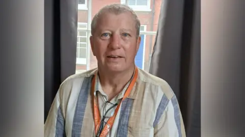 Alabaré A head and shoulder image of Alabaré Veteran Service Manager Mike Bedford with short grey hair and blue eyes wearing a beige, white a blue striped shirt, an orange lanyard and standing in front of a window with grey curtains.