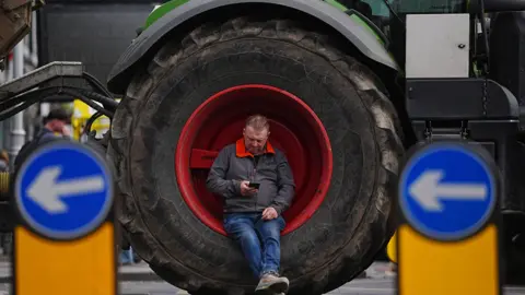 A man sits inside a large tractor wheel as vehicles are parked on O'Connell Street in Dublin. He's looking at his mobile phone. He's wearing a navy jacket with a red collar, blue jeans and brown boots.