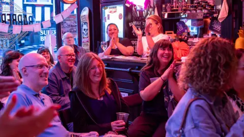A bar with pink lighting is filled with people sat down and laughing. The people watching are clapping and holding drinks. Two women are also seen standing behind a nearby bar. 