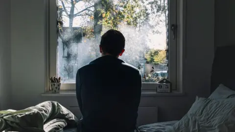 A man in front of a window, in a bedroom, in silhouette.