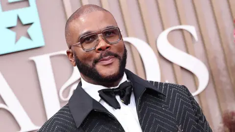 Tyler Perry is photographed on the red carpet of an event. He is wearing a black suit with a bow tie and is wearing square glasses. 