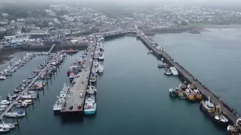 A drone shot of a harbour full of boats on a misty day. The water is a dark blue colour and there is mist and fog covering homes and the nearby town on the main land. 
