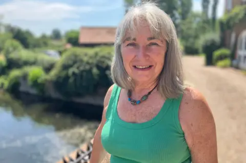 Owen Sennitt/BBC A woman wearing a green vest looks towards the camera while stood next to a bridge over a river, which is blurred in the background.