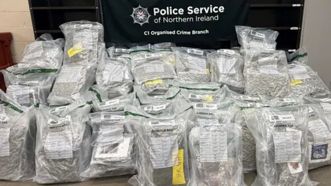 Blocks of green material can be seen inside over a dozen rucksack-sized plastic bags, which are lined and piled up in front of a banner reading "Police Service of Northern Ireland C1 Organised Crime Branch". 