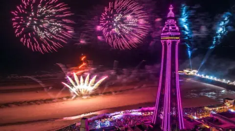 Blackpool Tower and the beach lit up in pinks and purples as fireworks blast against the night sky