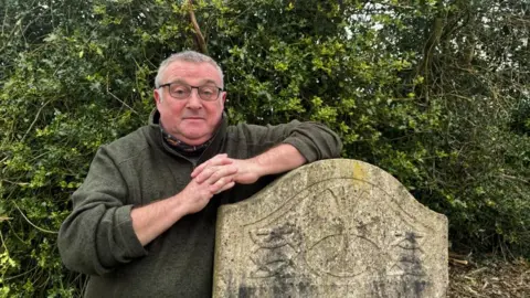 BBC/Alex James A man in a dark green quarter zip jumper is stood leaning against a grey, stained gravestone. Behind him is a green Holly bush