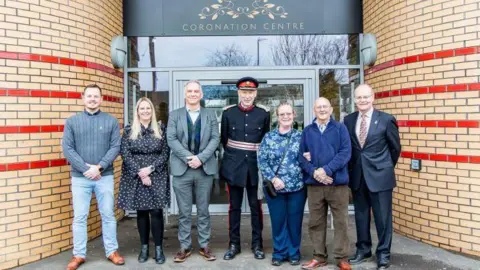 Six trustees of the Coronation Centre in Cadbury Heath stand with Vice-Lord Lieutenant of Gloucestershire Roger Deeks, who wears a military uniform, in front of the entrance to the newly refurbished building