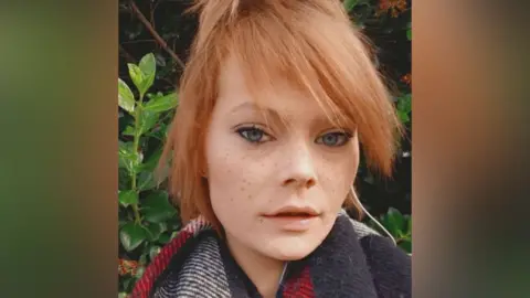 A selfie of a young woman, who has blue almond-shaped eyes, freckles and strawberry blonde hair, which is tied up. She is wearing a scarf and standing in front of greenery.