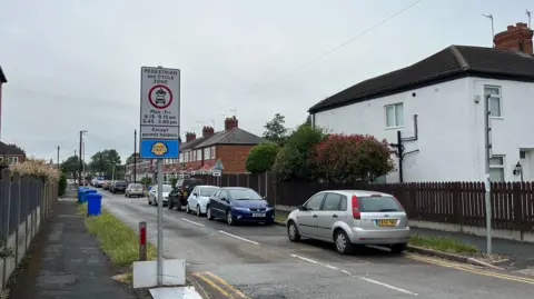 BBC/Richard Madden A row of cars parked on a residential street which is lined by a terrace of red brick houses. Two signs detailing a pedestrian and cycle zone are visible at the end of the street.