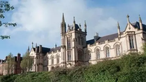 UU The university campus in Londonderry. A number of bushes and grassy areas can be seen in front of a Gothic-looking building with several large windows.
